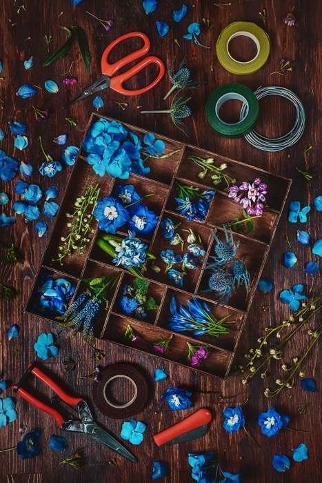 Flores de color azul cielo, pétalos y hojas de color rosa y violeta en una caja de madera, más pétalos y hojas sobre un fondo de mesa de madera, con tijeras, tijeras y cinta adhesiva