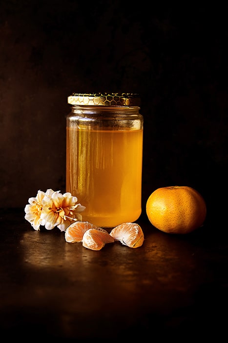 Foto de bodegón rústico de un tarro de miel, gajos de mandarina y flores sobre un fondo oscuro - asignaciones de fotografía