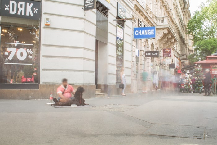 Una fotografía callejera de larga exposición en Budapest, un vagabundo borroso se sienta en el suelo con figuras borrosas en el fondo.