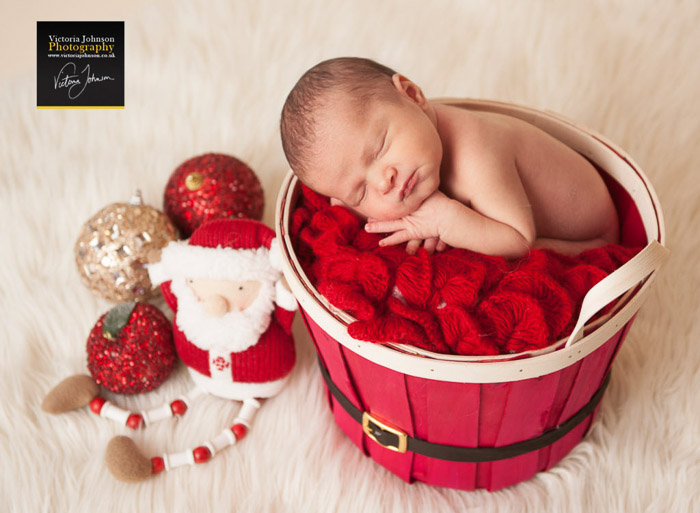 Un recién nacido en una canasta roja rodeado de accesorios de fotografía de recién nacidos con temática navideña