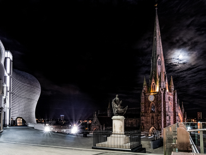 St Martin en Bullring, con la estatua de Nelson y el centro comercial Bullring en Birmingham.  La estructura iluminada contra un cielo oscuro, la luna espeluznante detrás de las nubes.