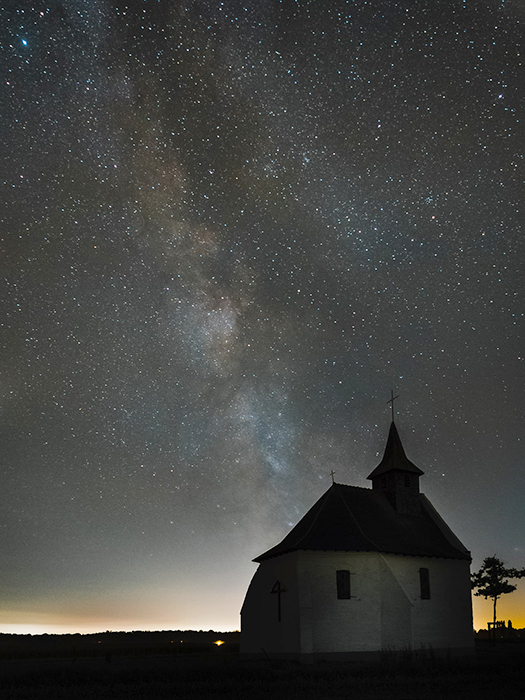 una capilla en una colina iluminada cálidamente desde atrás, contra el cielo nocturno con la vía láctea visible