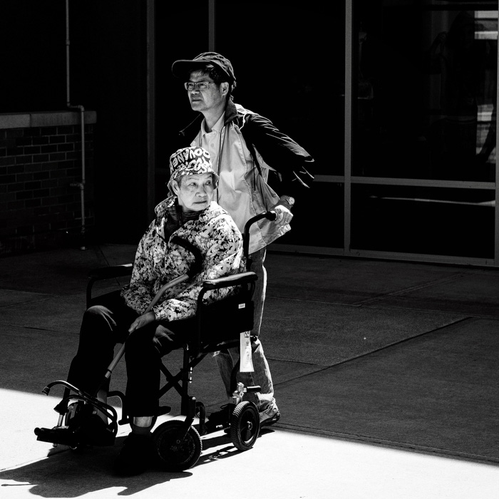 Un retrato en blanco y negro de un hombre empujando a una dama en una silla de ruedas al aire libre, filmado con procesamiento de película de extracción