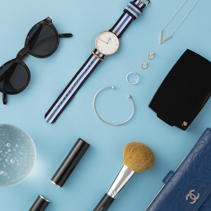 flatlay de accesorios de mujer sobre un fondo azul claro: reloj, pendientes, gafas de sol, cepillo, cartera