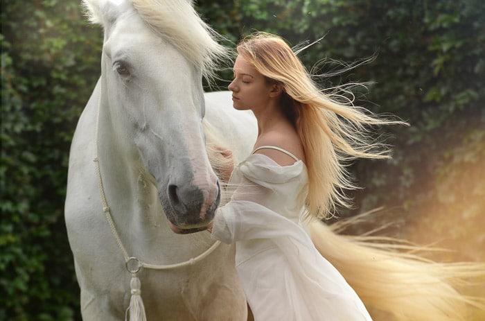 Una foto de ensueño de una modelo femenina con largo cabello rubio posando junto a un caballo blanco después de usar superposiciones de Photoshop 