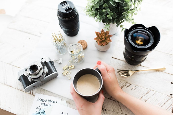 Fotografía cenital de una persona sosteniendo una taza de café en una mesa con plantas, cámara, lentes y otros equipos de fotografía