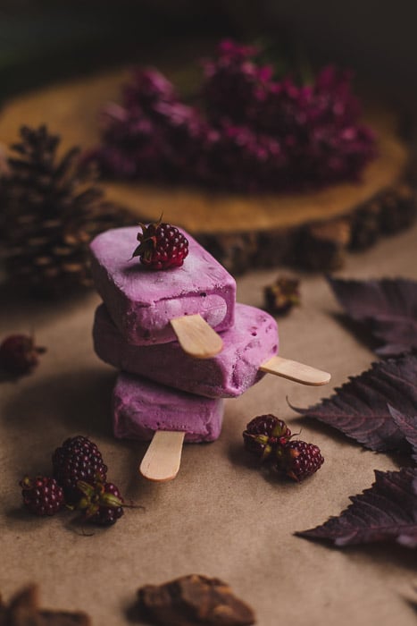 Una fotografía de alimentos oscura y cambiante toma de tres paletas de helado rosa en un montón sobre una superficie de madera con bayas y hojas