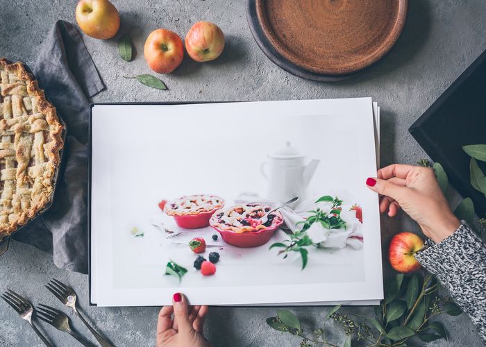 Manos sosteniendo impresiones fotográficas de comida sobre un escritorio con manzanas y un pastel