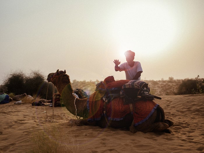 Un retrato de un hombre de pie junto a un camello en el desierto, con un hermoso efecto de destello de lente detrás de él.