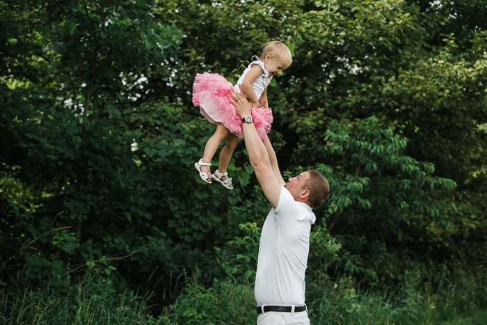Juguetón retrato de familia de un padre sosteniendo a una niña en tutú rosa sobre su cabeza en un frondoso escenario al aire libre