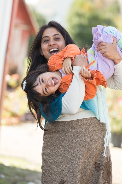 Retrato al aire libre de una madre sosteniendo a su hija juguetonamente boca abajo, ambos sonriendo hacia la cámara
