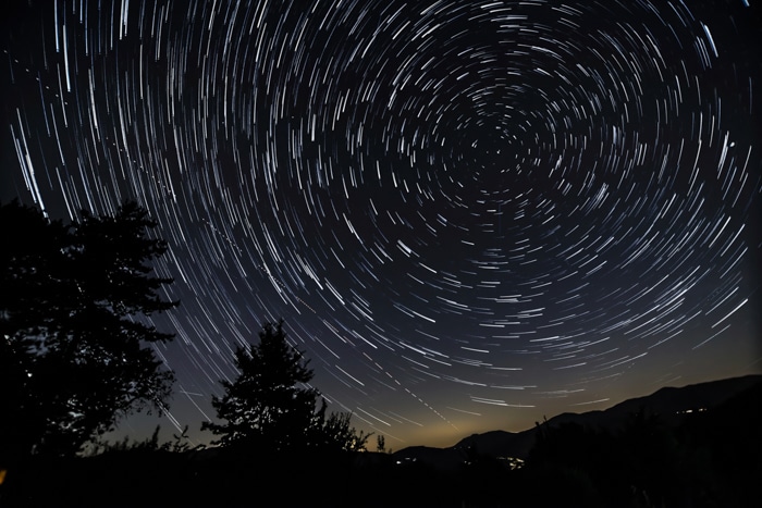 Fotografía de rastro de estrellas alrededor de la estrella del Polo Norte con colinas, árboles y luces de la casa.
