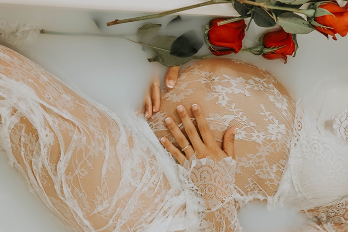 Una sesión de fotografía de baño de leche de cerca de una modelo de mujer embarazada con encaje blanco y sosteniendo su panza, rodeada de flores