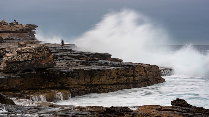 Foto de paisaje marino de dos personas pescando en las rocas con grandes olas detrás.