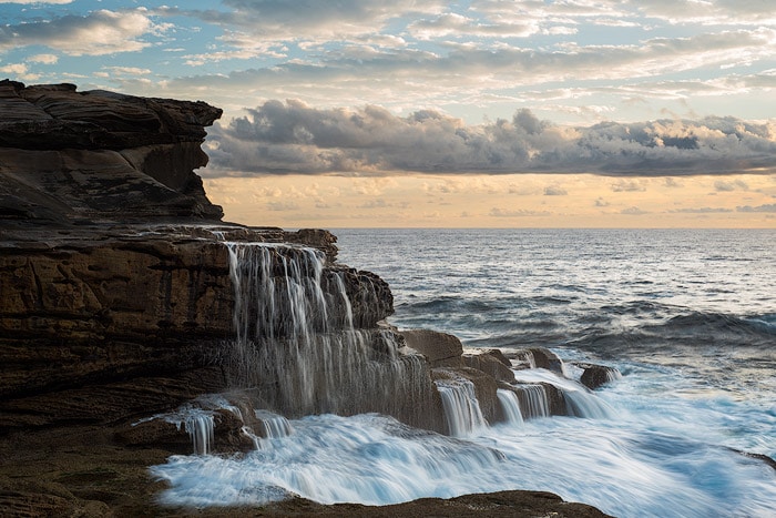 Foto de paisaje marino de una cascada corriendo hacia el océano, cielos nublados.