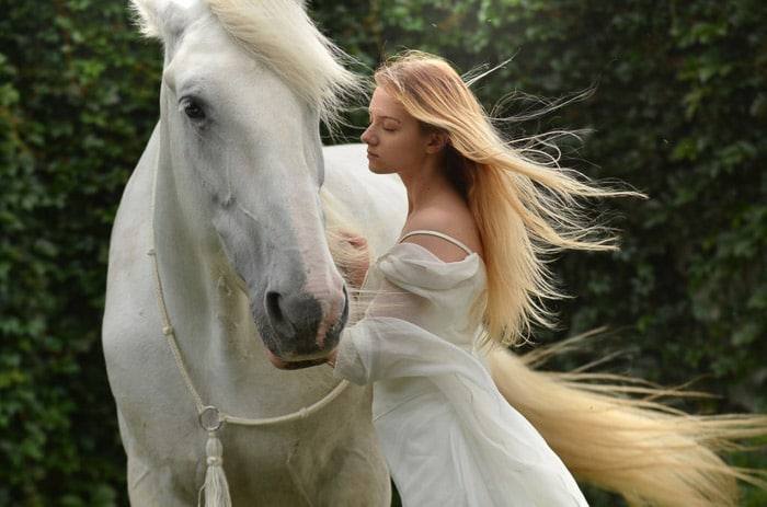 Una foto de ensueño de un modelo femenino con largo cabello rubio posando junto a un caballo blanco