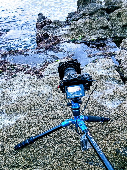 Imagen de una cámara y un trípode instalados en rocas costeras para fotografías de larga exposición.