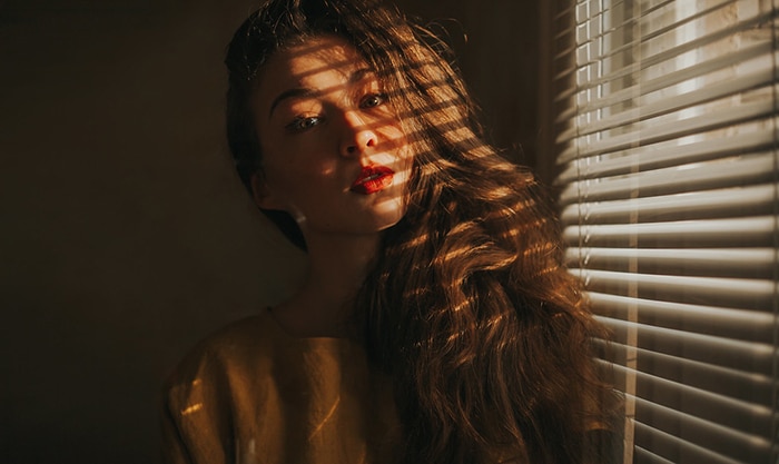 Un retrato temperamental de una modelo femenina posada junto a una ventana, las sombras de las persianas de las ventanas proyectadas en su rostro y cabello: ejemplos de retratos oscuros