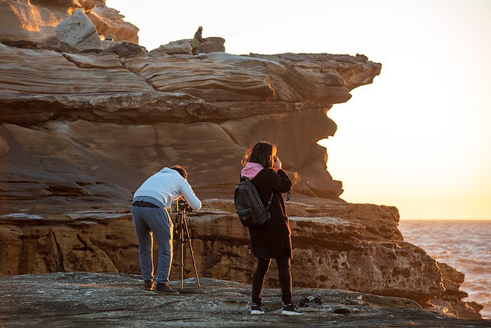 Hombre y mujer de pie en un acantilado rocoso, configurando una cámara y un trípode para fotografiar un paisaje marino