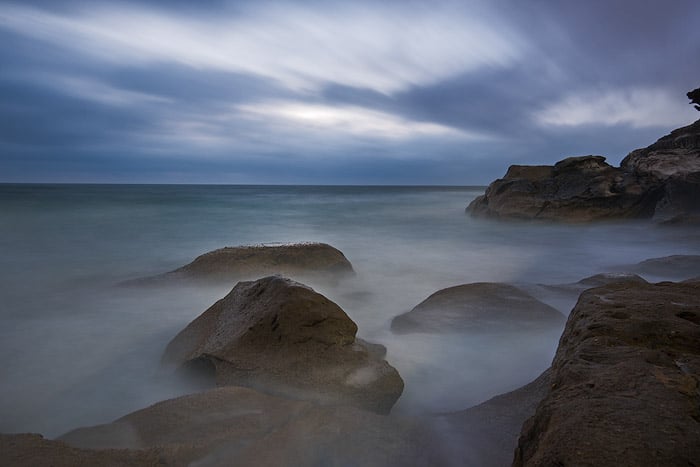 Una fotografía del océano de larga exposición toma de rocas que aparecen a través de un mar brumoso, un cielo nublado por encima
