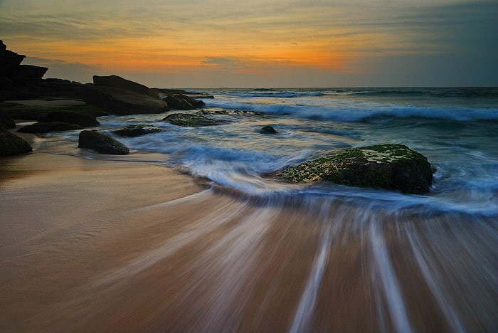 paisaje marino al atardecer.  senderos de agua en la orilla, rocas que aparecen en las olas espumosas, puesta de sol dorada en el horizonte