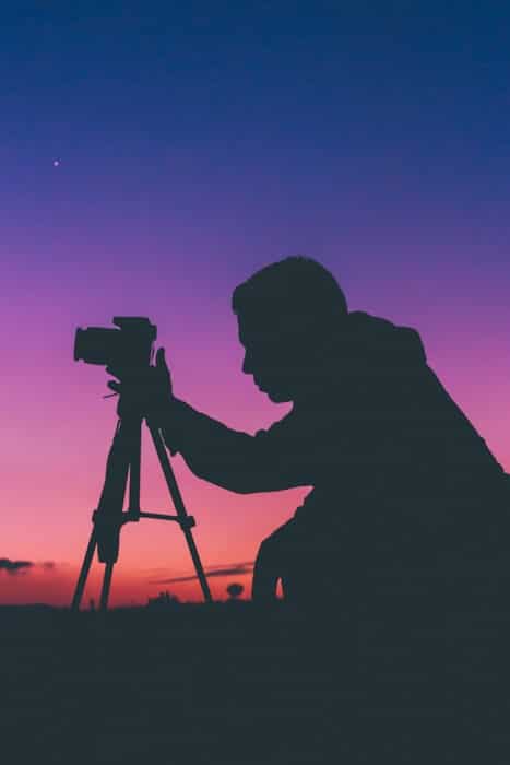 Silueta nocturna de un fotógrafo arrodillado ante su cámara sobre un trípode con un cielo de colores brillantes detrás de él
