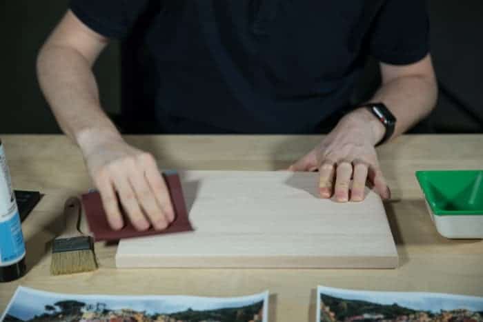 Un hombre se sienta en una mesa de madera, lijando una tabla de madera para preparar la superficie para transferir la foto a la madera