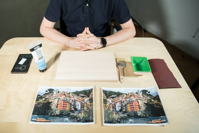 Un hombre se sienta en una mesa de madera, frente a él hay dos fotos idénticas de una ciudad costera de colores brillantes, pegamento, pincel, papel de lija y otros materiales para transferir la impresión láser a la madera.