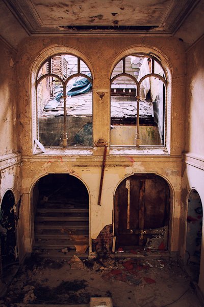 Edificio abandonado desde el interior, dos ventanas que dan a una azotea en ruinas y una escalera oscura que conduce hacia arriba.