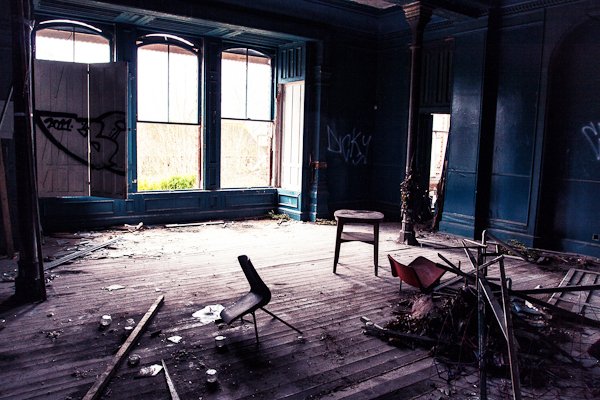 Habitación en un edificio abandonado con grandes ventanales, paredes pintadas de oscuro y madera y sillas rotas.