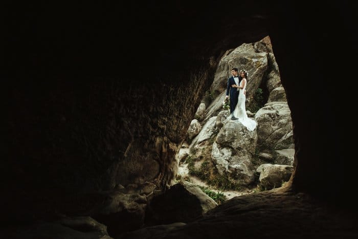 Una fotografía de boda de una pareja de pie fuera de la entrada de una cueva.