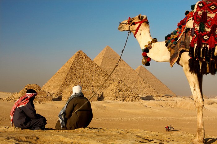 Dos beduinos descansando sobre la arena del desierto mirando las pirámides de Egipto mientras espera su camello