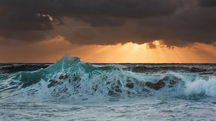olas espumosas rompiendo en la orilla, rayos dorados del sol asomando desde las nubes oscuras en el horizonte.  La velocidad del obturador es de 1/50 seg.