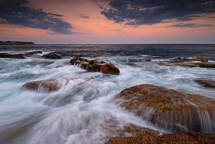 Aguas corriendo sobre rocas marrones planas en Coogee Beach, Sydney, Australia, un cielo anaranjado del atardecer en el horizonte