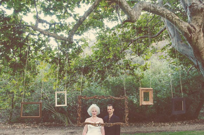 Un fotomatón de bodas genial instalado al aire libre con marcos de fotos de varios tamaños que cuelgan de una rama de árbol grande