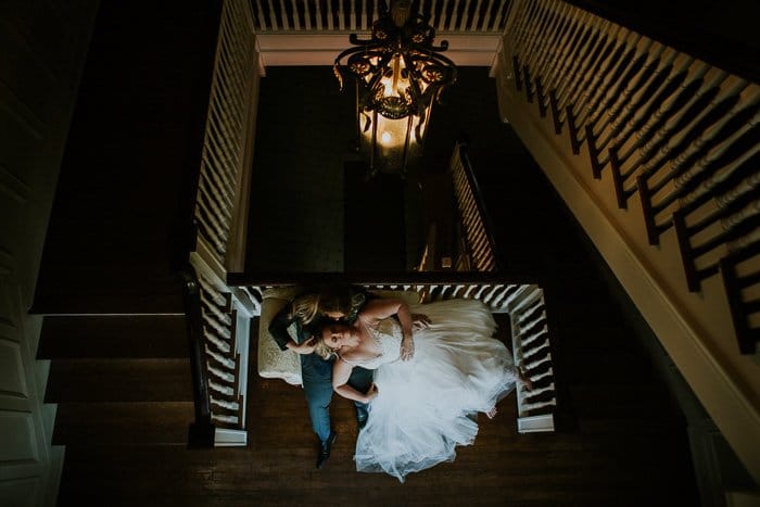 vista desde lo alto de las escaleras de una novia acostada en el regazo de su novio, en un sillón reclinable, la habitación iluminada por una lámpara de araña vintage