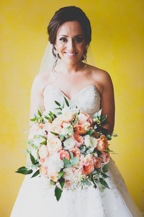 novia morena sonriente sosteniendo un ramo grande con una pared amarilla detrás de ella - tendencias de edición de fotos de bodas
