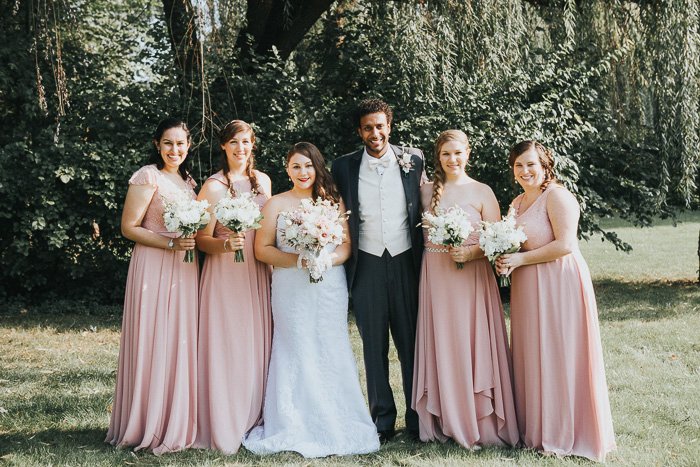 foto desaturada de la novia, el novio y las damas de honor en rosa pálido de pie contra un fondo de árboles.