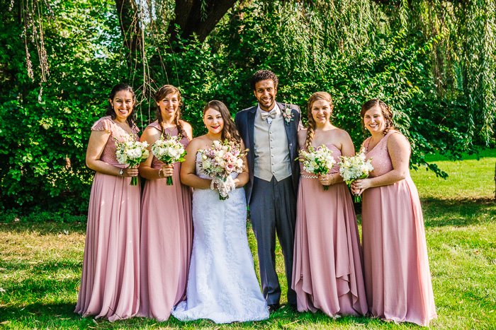 Foto HDR del séquito de bodas al aire libre contra árboles brillantes, novia, novio y damas de honor en rosa pálido