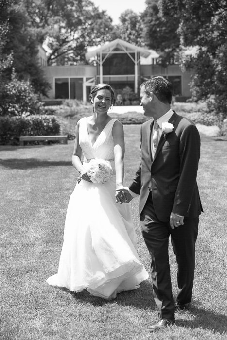 foto en blanco y negro de la novia y el novio recién casados ​​tomados de la mano, riendo, caminando por un césped