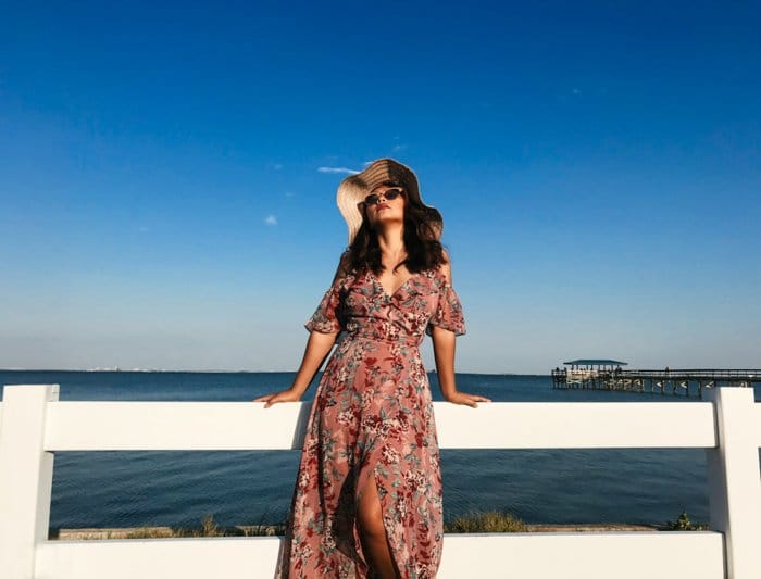 Mujer con un maxidress de verano, gafas de sol y un gran sombrero para el sol, apoyado en una valla blanca, el mar azul y el cielo detrás de ella