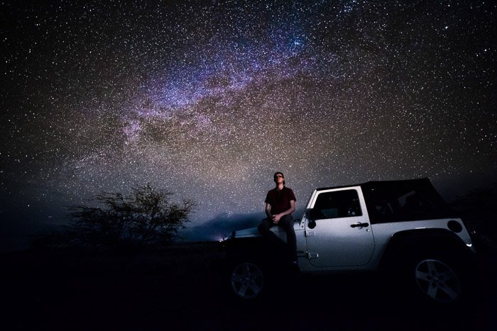 Toma de astrofotografía de fotograma completo de un hombre sentado en un jeep con un impresionante cielo lleno de estrellas detrás.  Tomada con lente rokinon de 14 mm a 30 segundos - F / 2.8 - ISO 3200