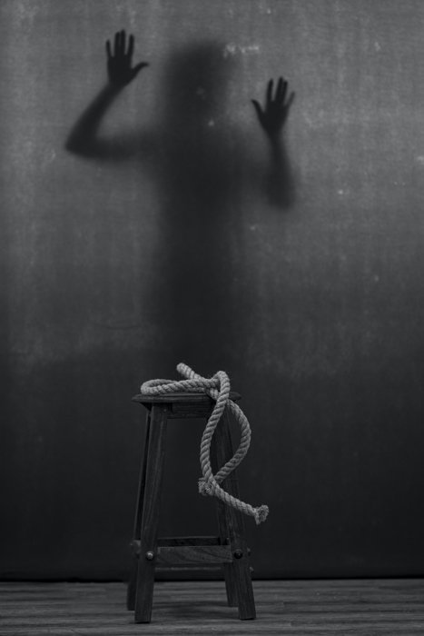 Imagen en blanco y negro de una cuerda en un taburete y una figura sombría en el fondo