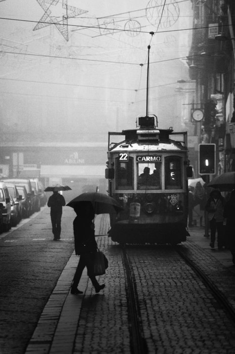 Imagen en blanco y negro de un hombre con paraguas cruzando un tranvía