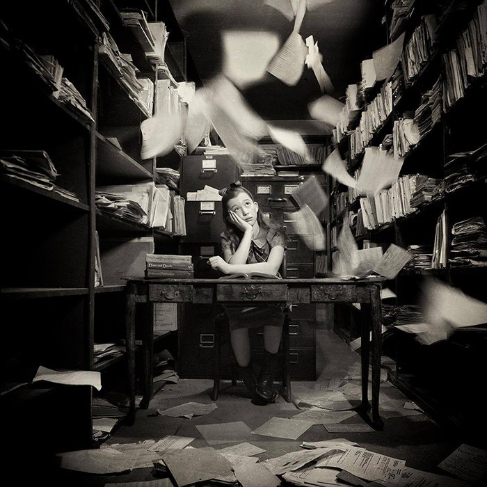 Conceptual retrato en blanco y negro de una joven sentada en una oficina con papeles volando por la fotógrafa de bellas artes Carolyn Hampton