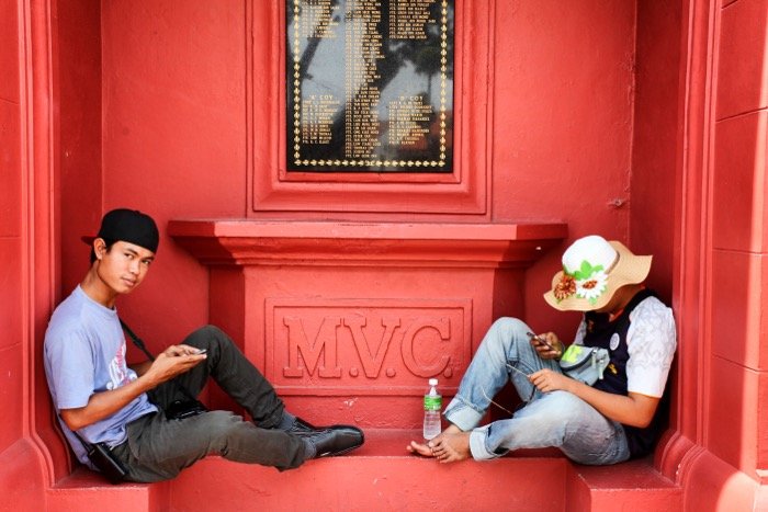 Fotografía callejera de dos personas sentadas en una pared roja.