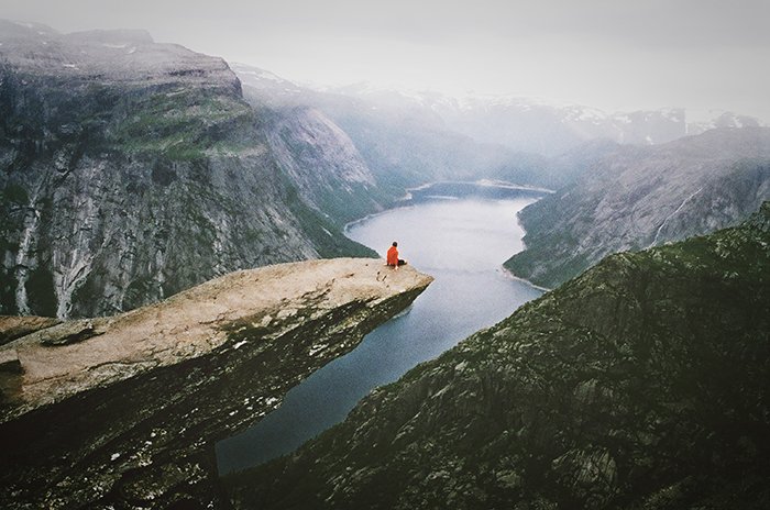 Un ejemplo de fotografía sincera de una persona sentada en un acantilado con vistas a un impresionante paisaje montañoso. 