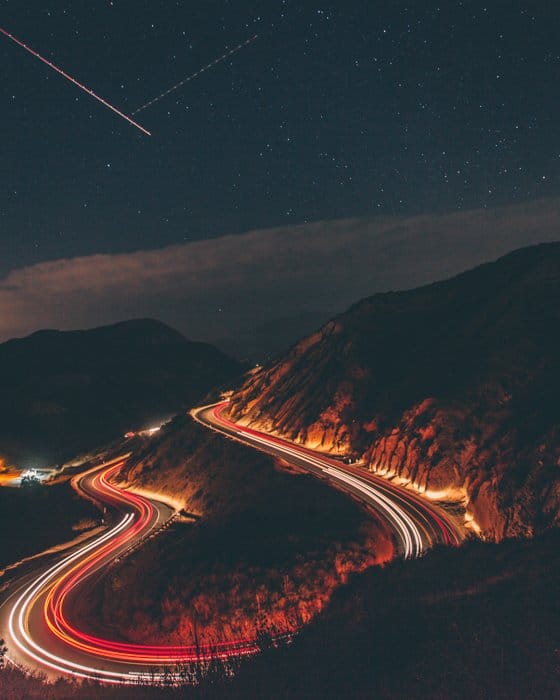 Una foto de lapso de tiempo de automóviles conduciendo por una carretera de montaña curva con estrellas fugaces en el cielo