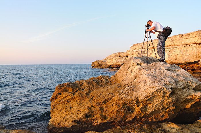 Hombre de pie sobre una roca y tomando fotos con trípode en hora azul.