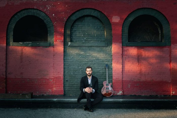 Un retrato en color de un músico posando delante de una pared roja en Brooklyn
